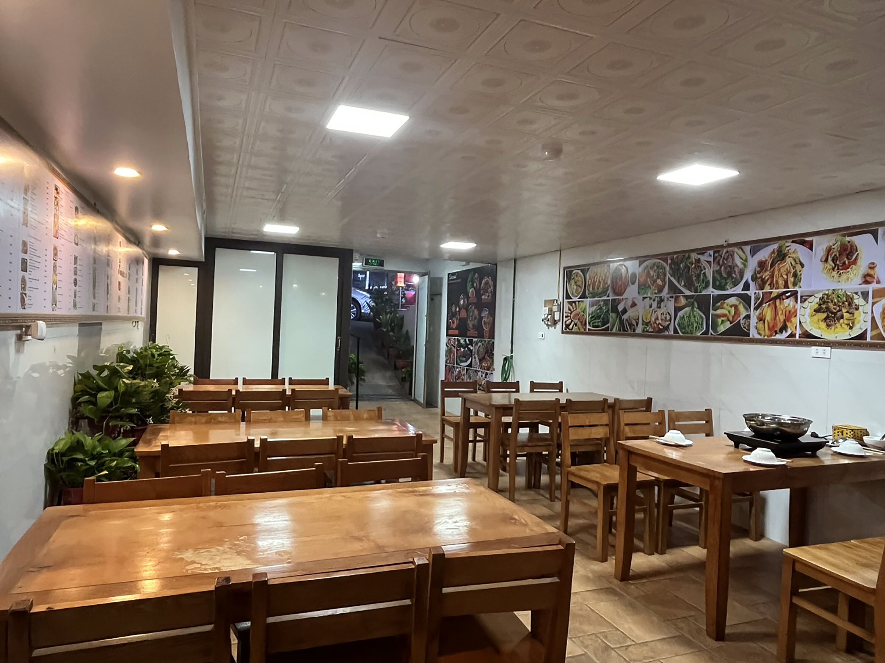 A restaurant interior with wooden tables and chairs, a menu board on the left, and a wall of food images on the right.