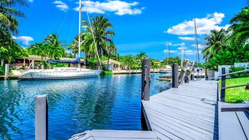 A wooden dock extending into a calm waterway with boats, surrounded by lush greenery and palm trees.