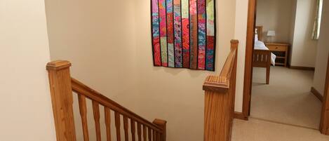 A wooden staircase with a handrail, carpeted floor, and a colorful quilt hanging on the wall.