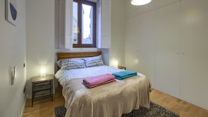 Un dormitorio con cama, mesita de noche, lámpara, ventana con cortinas y armario.