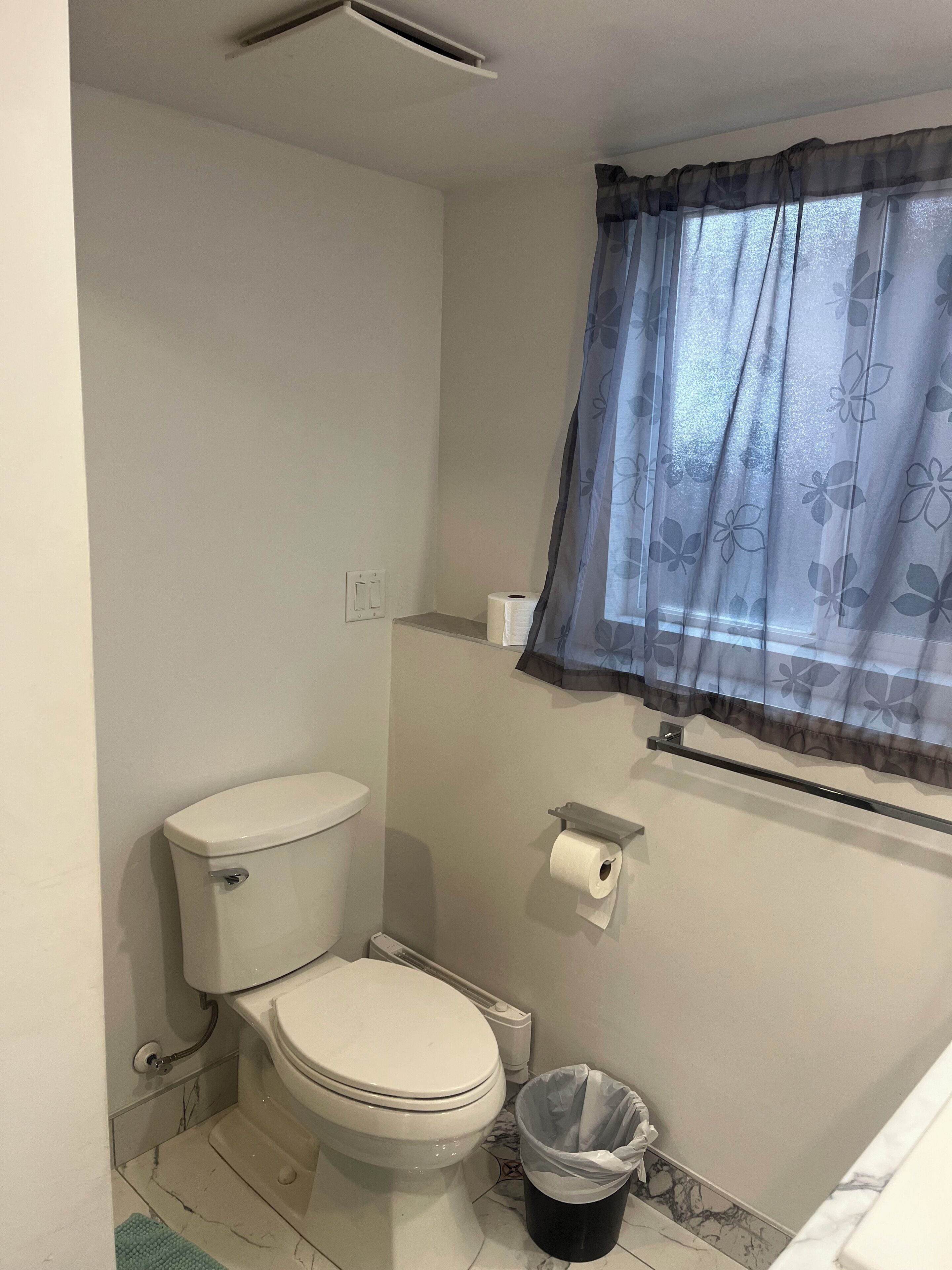 Un bagno piccolo e pulito con il water, un cestino, un porta carta igienica e una finestra con tenda a fiori.
