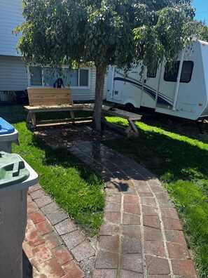 A paved pathway, a wooden bench, a tree, and a white RV parked in a grassy area.