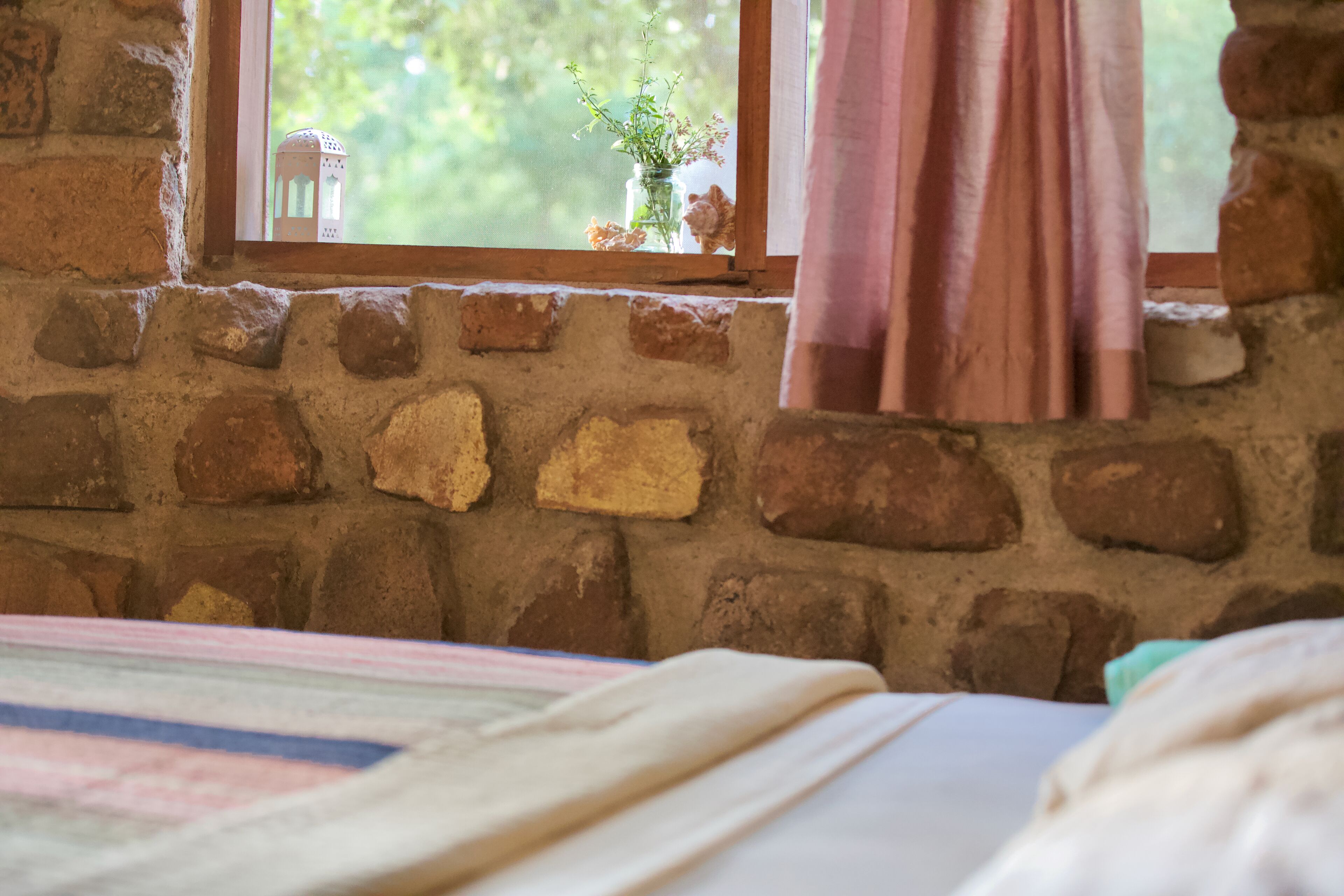 Una habitación rústica con pared de ladrillo, ventana con vistas a follaje y cama con manta a rayas.