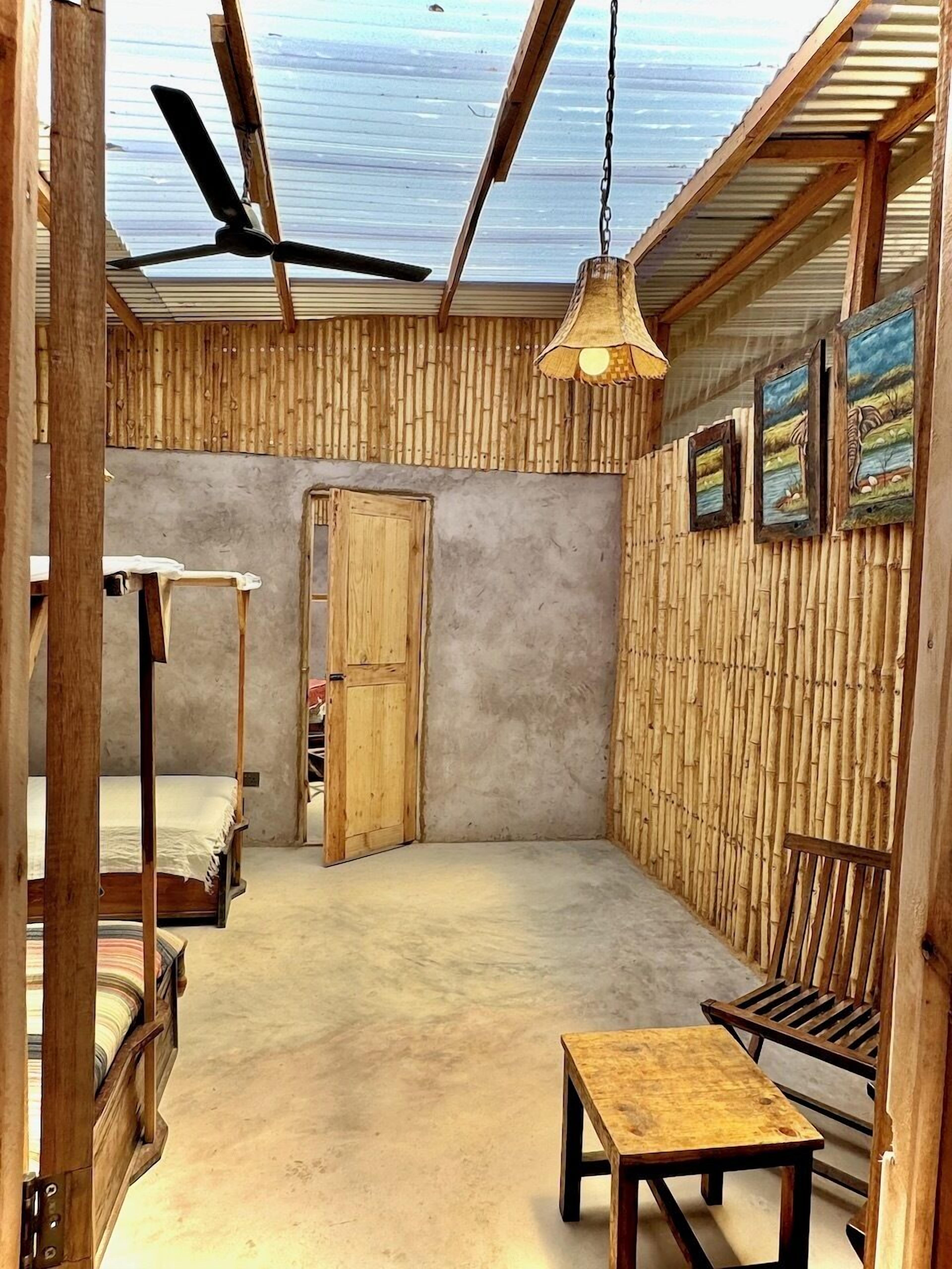 A room with a bunk bed, a table, a bench, a ceiling fan, and bamboo wall panels.