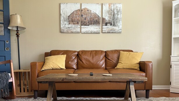 Een bruine leren bank met twee gele kussens, een houten salontafel en een schilderij van een schuur aan de muur.