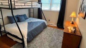 Een stapelbed met grijs-witte beddengoed, een klein nachtkastje met een lamp, en een raam met gordijnen.