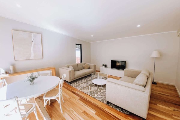A bright living space with two gray sofas, a flat-screen TV, a round glass coffee table, and a nearby dining area. The room features wooden floors, wall art, and ample natural light for a cozy feel.