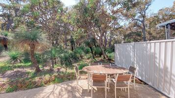 An outdoor patio with a round wooden table and chairs, surrounded by a white fence and various trees.