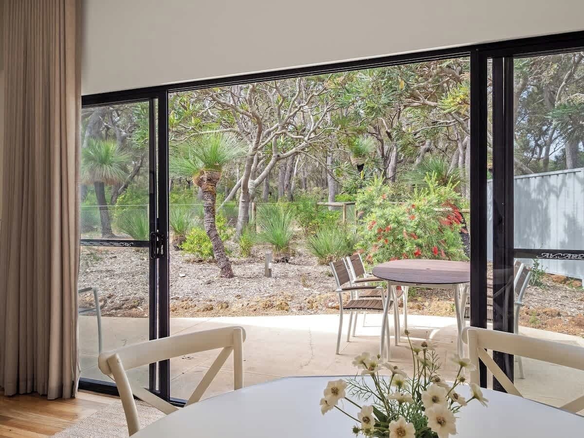 Habitació amb una gran porta corredissa de vidre que dona a un pati exterior amb taula i cadires, envoltat de vegetació i arbres.