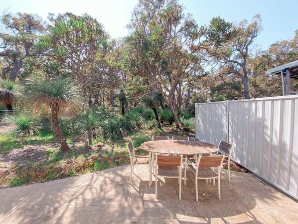 An outdoor patio with a round table and chairs, surrounded by a white fence and various trees.