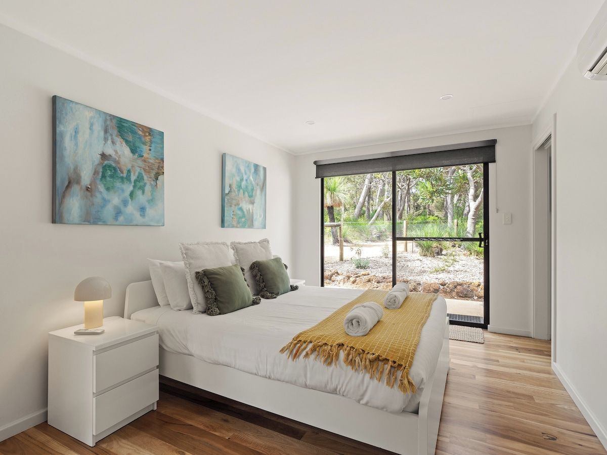 A modern bedroom with a large bed, a bedside table, and a view of the outdoors through a sliding door.