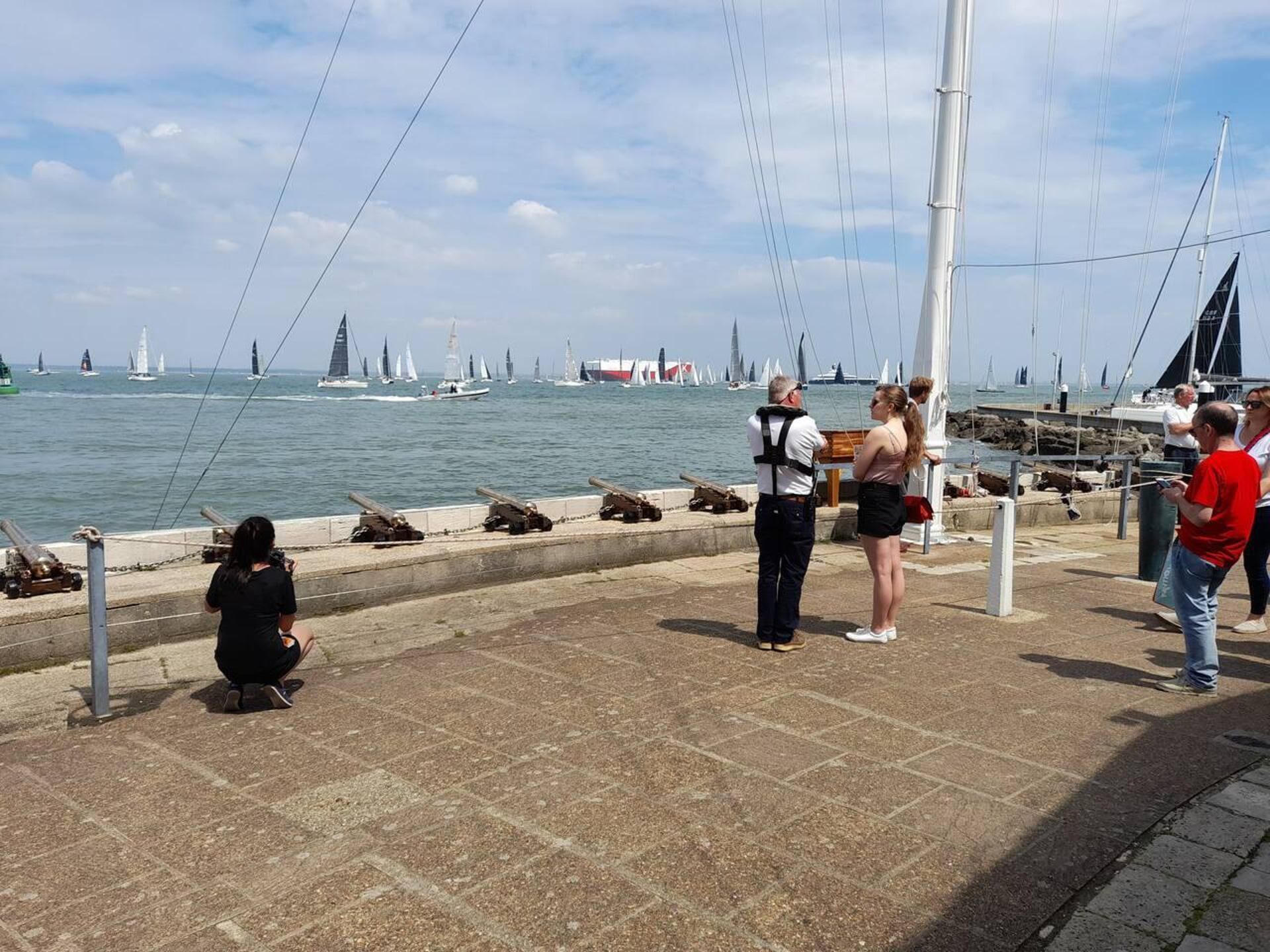 A waterfront promenade with a row of cannons, a pier with boats, and a clear sky.