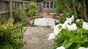 Un jardin avec une terrasse en pierre, une clôture en bois et un abri de jardin à l’arrière-plan.
