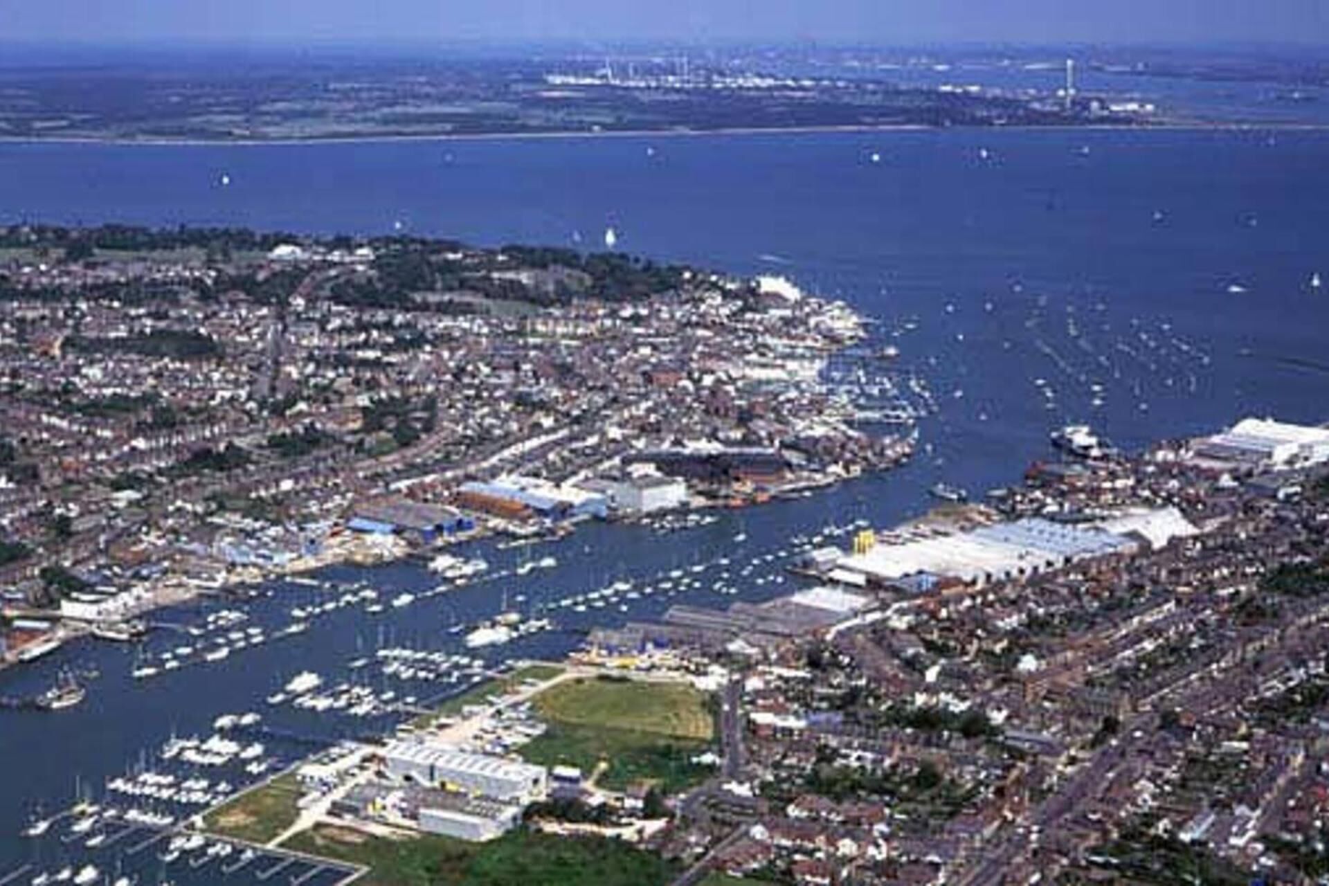 An aerial view of a coastal city with dense urban development, a marina with numerous boats, and a large industrial area.