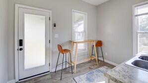 Una cocina con una mesa de madera y dos taburetes altos, una puerta con mirilla y una ventana con persianas.