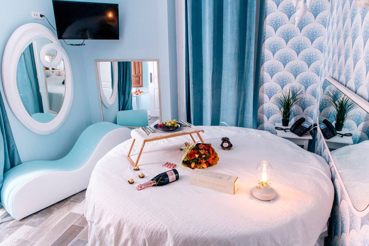 Un dormitorio moderno con una cama curvada, un espejo grande, un televisor de pantalla plana y una mesita de noche con una lámpara y un libro.
