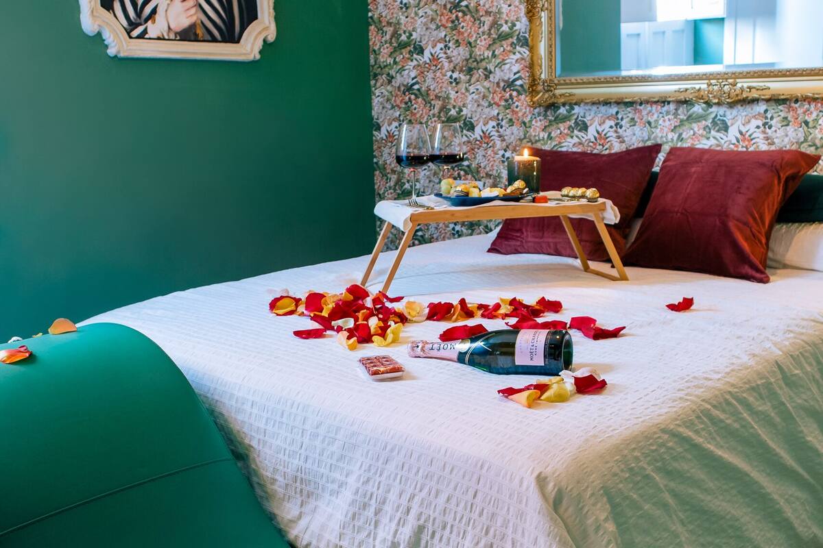Un dormitorio con pared verde, papel tapiz floral, un cuadro con temática de cebra, cama con pétalos de rosa, una botella y una bandeja con velas.