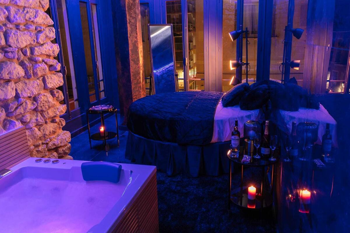Una habitación tenuemente iluminada con pared de piedra, una cama grande, un jacuzzi y una mesa con velas y botellas.