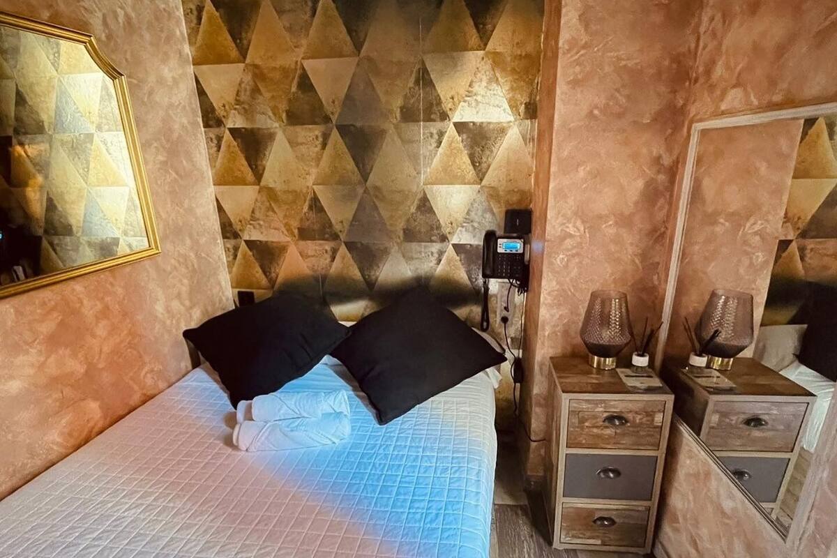 Un dormitorio con pared de estampado geométrico, una cama con almohadas negras, una mesita de noche con lámpara y un espejo.