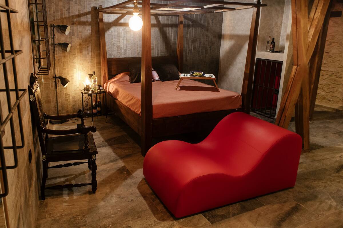 Una habitación con cama de dosel, un sofá rojo, una silla de madera, una mesita con una botella y vasos, y una estantería de pared con una botella.