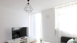 Ein modernes Wohnzimmer mit einem Flachbildfernseher auf einer weißen Kommode, einem Couchtisch mit einer Vase, einem grauen Sofa und einem Fenster mit Vorhängen.