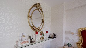 Una habitación con un espejo grande y decorativo, un estante de vidrio con artículos de tocador, una mesita redonda con una planta y una cama con cabecera roja.
