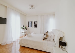 A bedroom with a large bed, a bedside table with a lamp, a desk with a chair, and a wall-mounted TV.