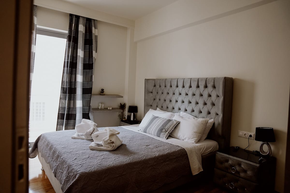 Una cama bien hecha con cabecera gris, una mesita de noche con lámpara, y una ventana con cortinas.