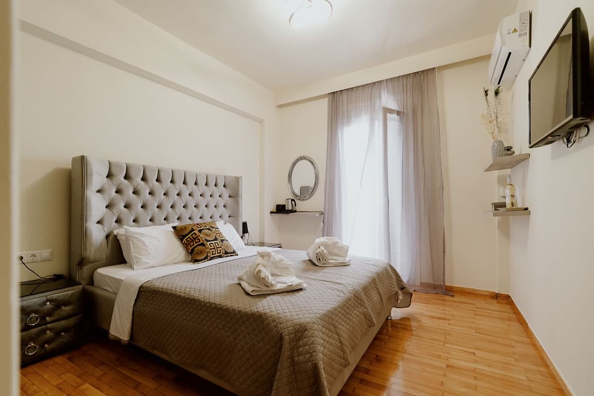 Un dormitorio ordenado con una cabecera capitonada, una cama con cubrecama acolchado, dos toallas sobre la cama, una mesita de noche, un espejo y un televisor montado en la pared.
