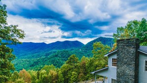 Une maison avec une cheminée en pierre, un porche et une vue sur une chaîne de montagnes boisées.