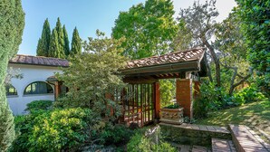 Un jardín con un pérgola, un edificio blanco con ventanas de arco y abundante vegetación.