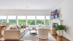 Un salon spacieux avec un canapé beige, une table basse ronde et un fauteuil blanc. De grandes fenêtres offrent une vue sur la verdure environnante.
