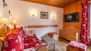 Ein gemütliches Zimmer mit Holzbalkendecke, einer Couch mit gemusterten Kissen, einem kleinen Tisch, einem Fernseher und einem roten Vorhang.