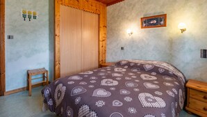 Ein Schlafzimmer mit Holzbalkendecke, einem Bett mit gemustertem Überwurf, einem Nachttisch aus Holz, einem kleinen Holzschemel und einer Wand mit einem gerahmten Bild und Wandleuchten.