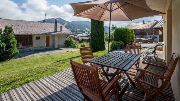 Eine Holzterrasse mit Tisch und Stühlen, einem Sonnenschirm und Blick auf Häuser und Berge im Hintergrund.