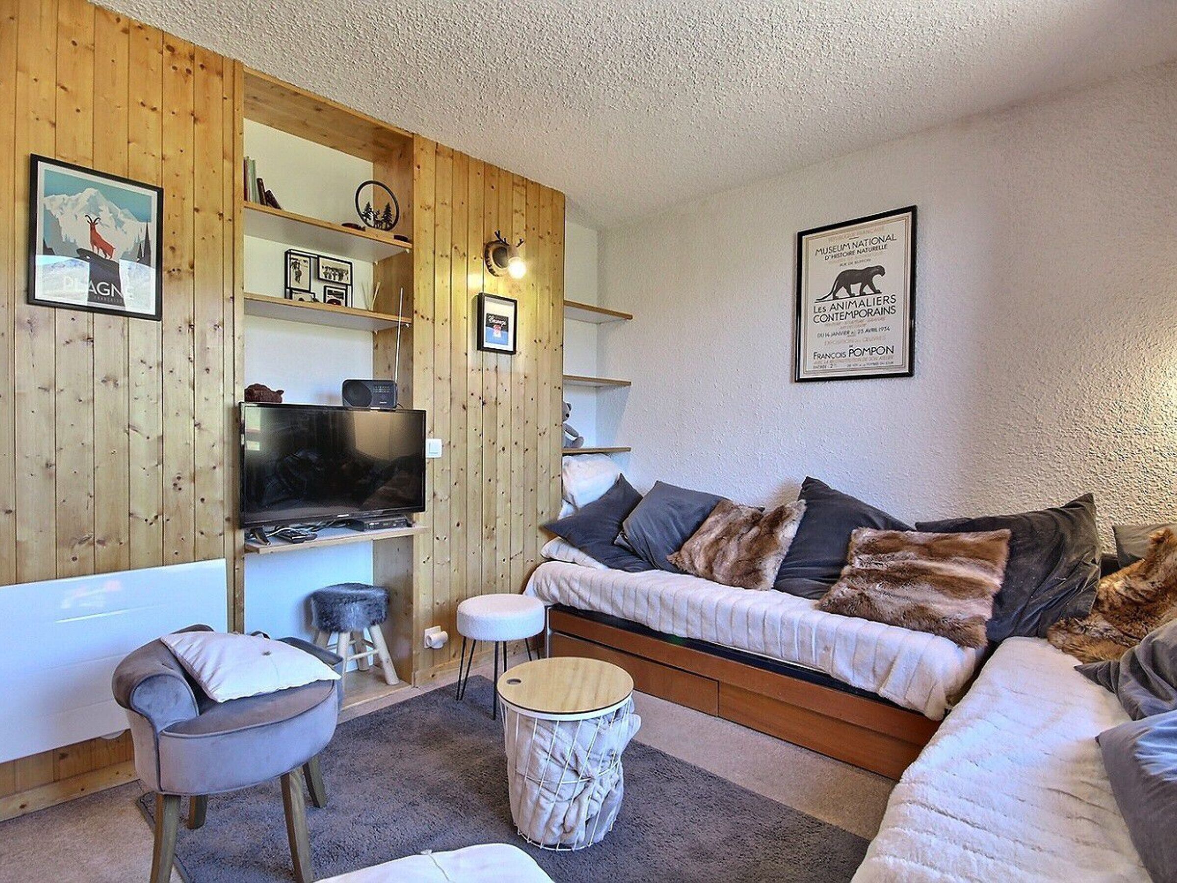 A cozy living room with a wooden wall, a flat-screen TV, a sofa with cushions, a small stool, and a round table.