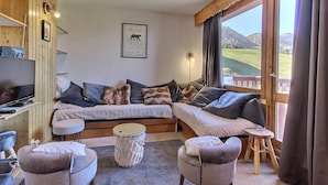 Una sala de estar acogedora con un sofá, dos sillones, una mesita redonda y un televisor. Un cuadro enmarcado cuelga de la pared, y una ventana ofrece vistas a un paisaje montañoso.