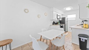 Une cuisine compacte avec des meubles blancs, une table à manger avec des chaises, une petite poubelle et un grille-pain jaune.