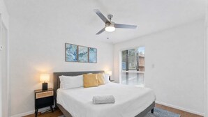 Une chambre à coucher avec un lit, des tables de chevet, un ventilateur de plafond, des œuvres d’art et une fenêtre.