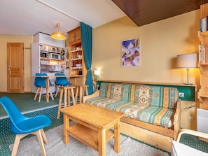 A cozy living room with a sofa, chairs, a small wooden table, a kitchen area, and a bookshelf.