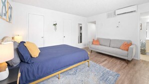 A bedroom with a bed, a sofa, a nightstand, a lamp, a wall-mounted air conditioner, and a door. - Central Hyde Park studio close to UT, Moody Center (Austin)