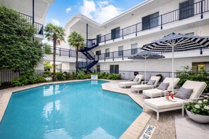 A swimming pool with lounge chairs and umbrellas, surrounded by greenery and a multi-story building with balconies.