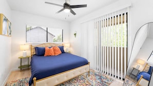 Un dormitorio con una cama, dos mesitas de noche, un ventilador de techo, una ventana con persianas y una alfombra estampada.