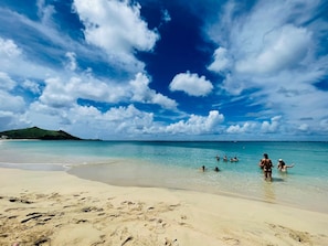 A beach with clear turquoise water, sandy shore, and a distant green hill.