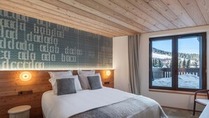 A cozy bedroom with a wooden ceiling, a large bed with white and gray bedding, a wall-mounted light, and a window with a snowy view. - New 86m² in Courchevel 1850 - Ski-in/Ski-out, for 4 adults and 2 kids (Courchevel 1850)
