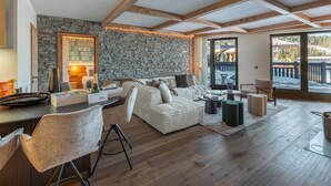 A cozy living room with a stone wall, wooden beams, a sofa, a coffee table, and a view of a snowy landscape through a glass door. - New 86m² in Courchevel 1850 - Ski-in/Ski-out, for 4 adults and 2 kids (Courchevel 1850)