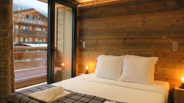 Hotellihuone, jossa on puuseinä, valkoisilla lakanoiden peitetty sänky, ruutukuviollinen peitto ja ikkuna, josta avautuu näkymä rakennukseen, jonka kyltissä lukee ”Chamois d’Or”.