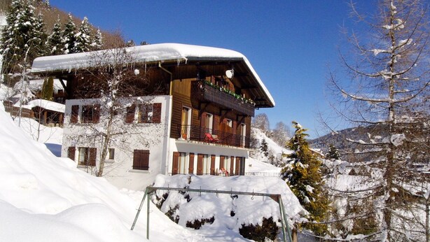 A multi-story chalet with a snow-covered roof and balcony, surrounded by snow-covered trees and a clear sky.