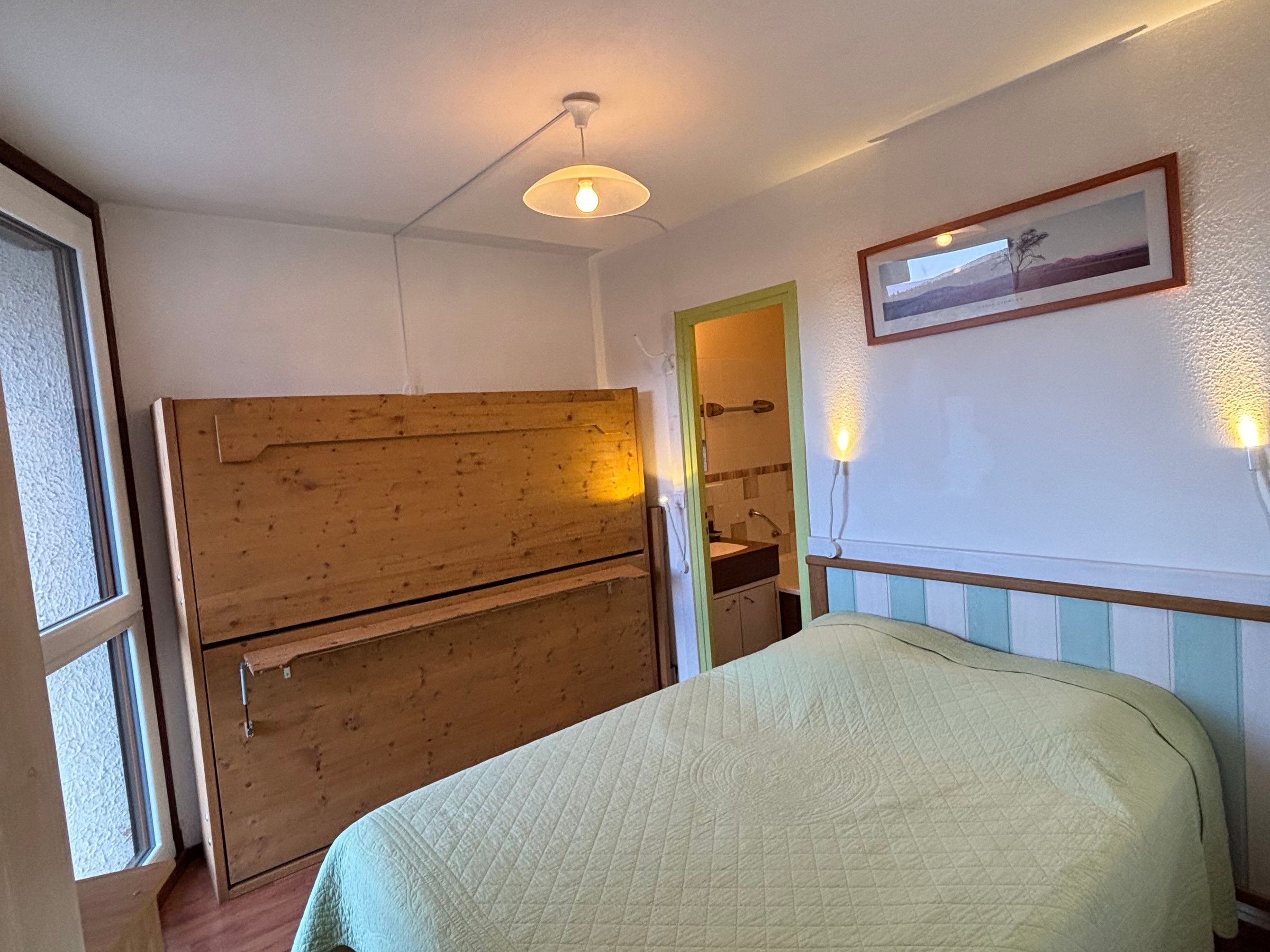Ein Zimmer mit einem Bett, einem Holzkoffer, einem gerahmten Bild an der Wand und einem Fenster.