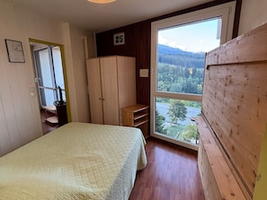 A bedroom with a bed, a wardrobe, a desk, and a large window with a view of a forest and mountains.